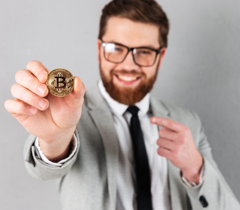Portrait of a satisfied businessman Portrait of a satisfied businessman dressed in suit pointing finger at bitcoin and looking at camera isolated over gray background